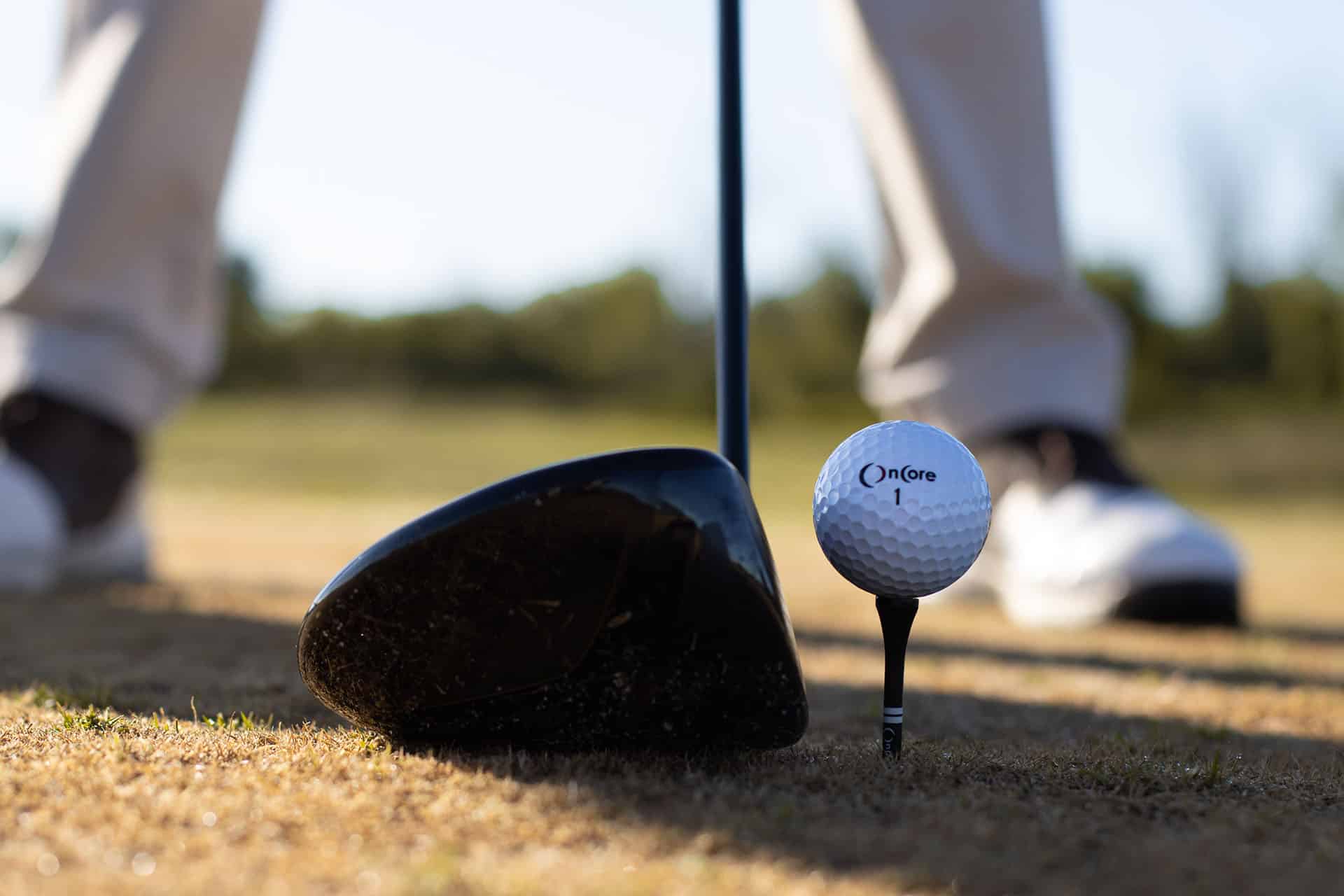 A close-up view of a golf club resting beside a golf ball on a tee, with the legs of a golfer visible in the background, capturing the preparation for a golf swing.