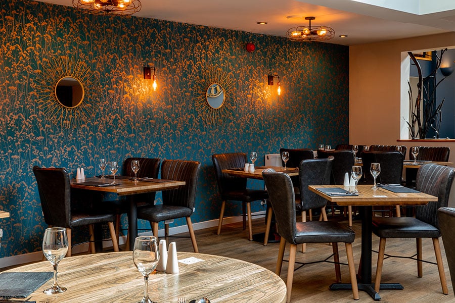 An interior view of a restaurant adorned with a decorative wallpapered wall. The dining area is neatly set with tables and chairs, though currently unoccupied.