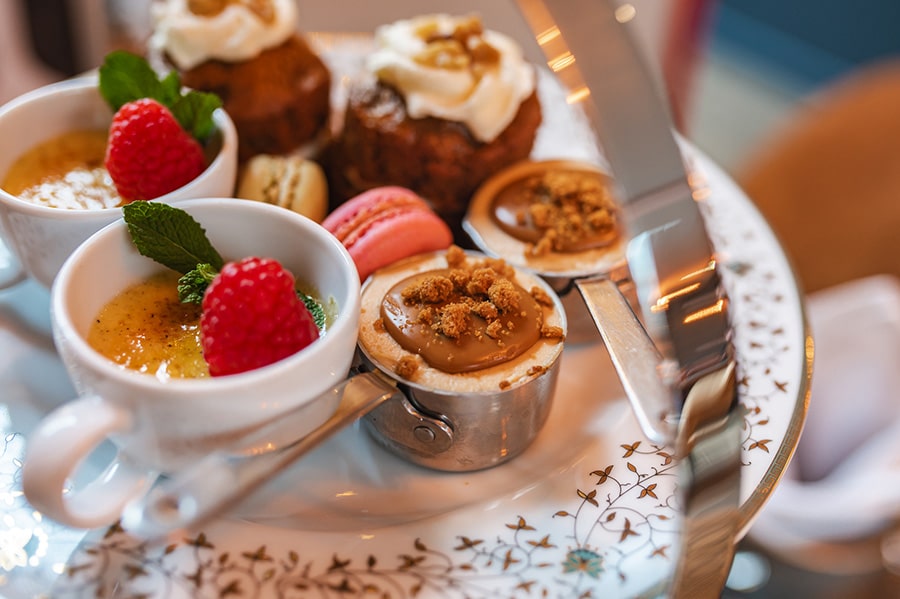 An elegant afternoon tea selection featuring crème brûlée with raspberries, caramel mousse cups with biscuit crumbs, macarons, and walnut-topped cakes on a decorative tiered tray.