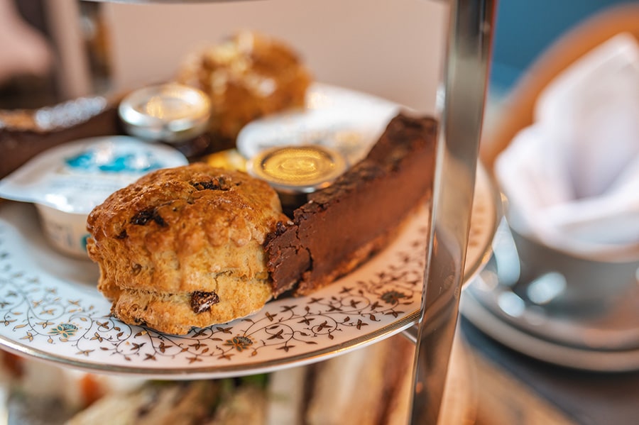 A close-up of an afternoon tea stand featuring a golden raisin scone, a rich chocolate tart, clotted cream, and jam on an elegant floral-patterned plate.