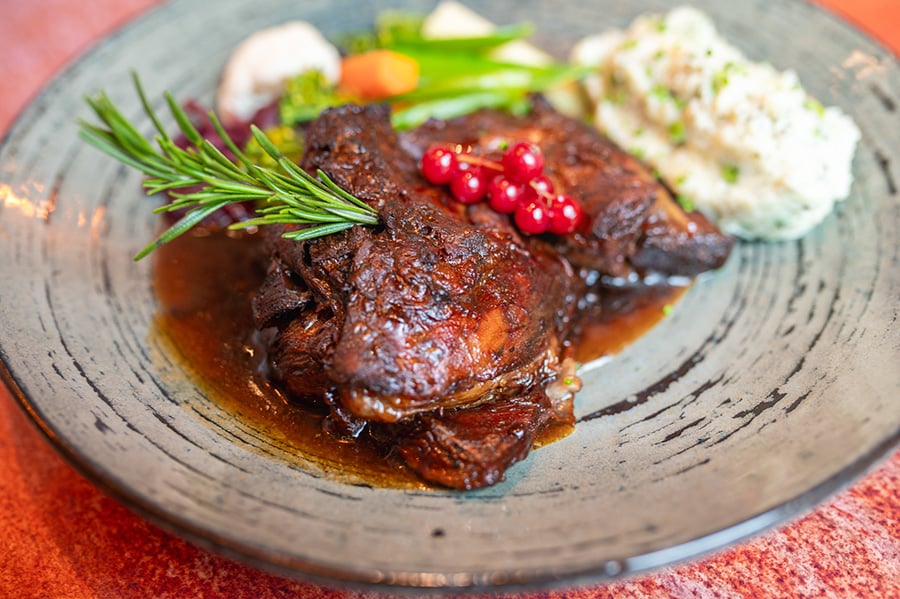 A dish of slow-braised beef in a rich sauce, served with creamy mashed potatoes, fresh vegetables, and garnished with redcurrants and rosemary on a rustic ceramic plate.