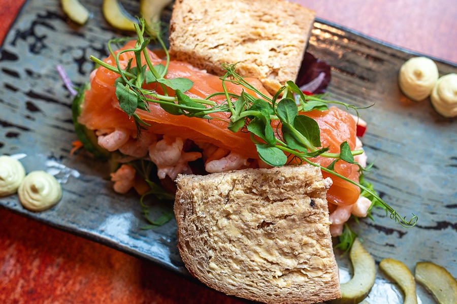A smoked salmon and prawn sandwich served on rustic wholemeal bread, garnished with fresh pea shoots and accompanied by pickles and a creamy sauce, presented on a ceramic plate.