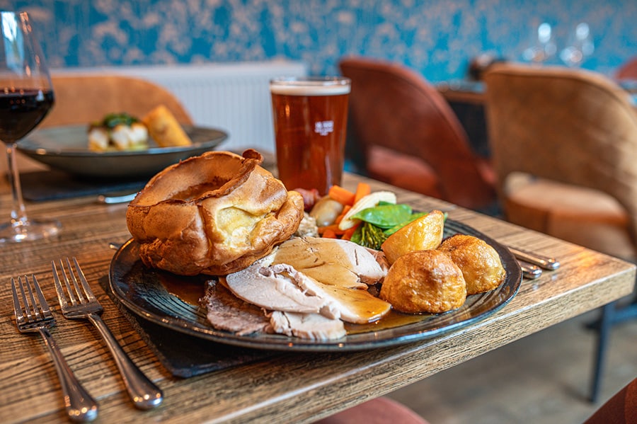 A classic Sunday roast dinner featuring slices of roast turkey and beef, golden roast potatoes, Yorkshire pudding, seasonal vegetables, stuffing, and gravy, served on a rustic plate in a cosy restaurant setting. A pint of beer and a glass of red wine are visible in the background.