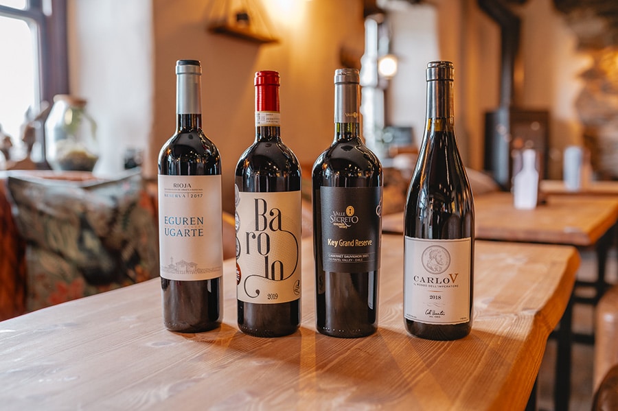 A selection of four red wine bottles, including Rioja, Barolo, Cabernet Sauvignon, and Carlo V, elegantly displayed on a wooden table in a cozy bar setting.
