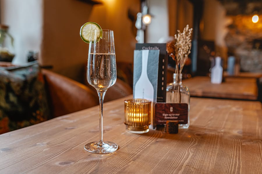 A glass of sparkling wine with a lime garnish on a rustic wooden table, set in a cozy bar with warm lighting and elegant decor.