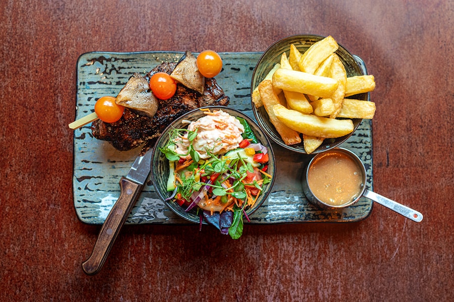 Grilled steak with skewered mushrooms and cherry tomatoes, served with thick-cut chips, fresh salad, and a side of sauce on a rustic plate.