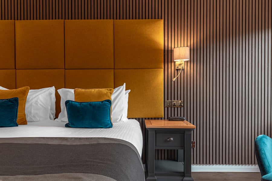 Modern hotel room with a mustard yellow upholstered headboard, decorative pillows, and a stylish wooden bedside table.