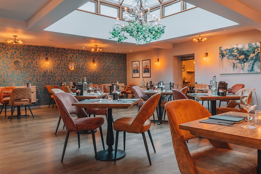 Stylish restaurant interior with leather chairs, wooden tables, warm lighting, and a decorative chandelier beneath a skylight.